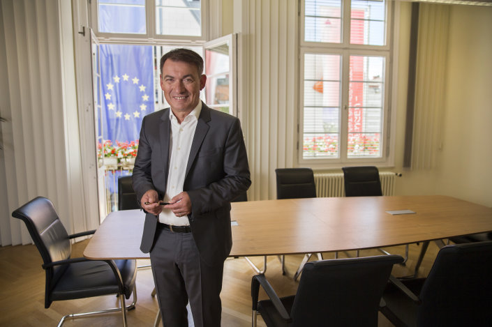 Guido Till steht in einem Raum vor einem Besprechungstisch, durch die göffneten Fenster sieht man die Flagge der EU