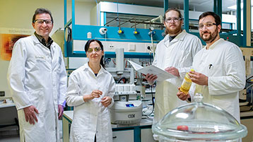 Prof. Dr. Ulrich Schörken, Devrim Eren Seitz, Lars Nitzsche und Tristan Jolmes stehen, mit weißen Laborkitteln bekleidet, in einem Labor (Bild: Michael Bause/TH Köln)