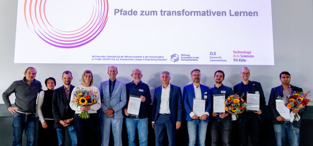 Elf Personen stehen auf der Bühne, einige halten einen Blumenstrauß oder einen Preis. Über ihnen eine Präsentation:  Elf Personen stehen auf der Bühne, einige halten einen Blumenstrauß oder einen Preis. Über ihnen eine Präsentation: