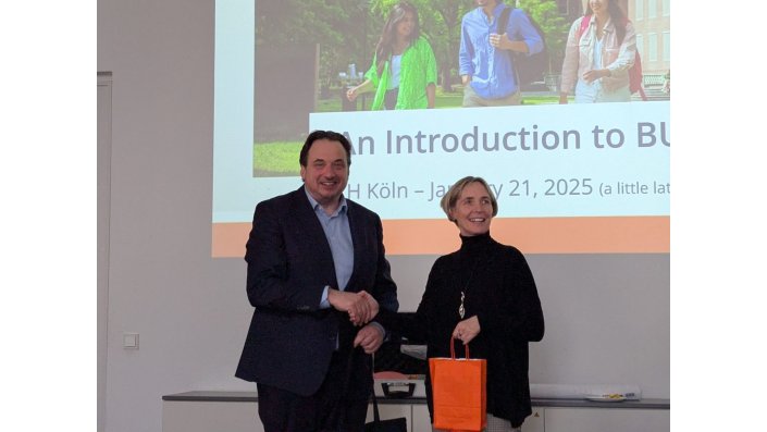 Dr. Jorrit Snyder on the left, wearing a dark blue jacket and a light blue dress shirt, present a gift bag to Prof. Dr, Sylvia Heuchemer, who wears a black turtleneck. They are shaking hands.