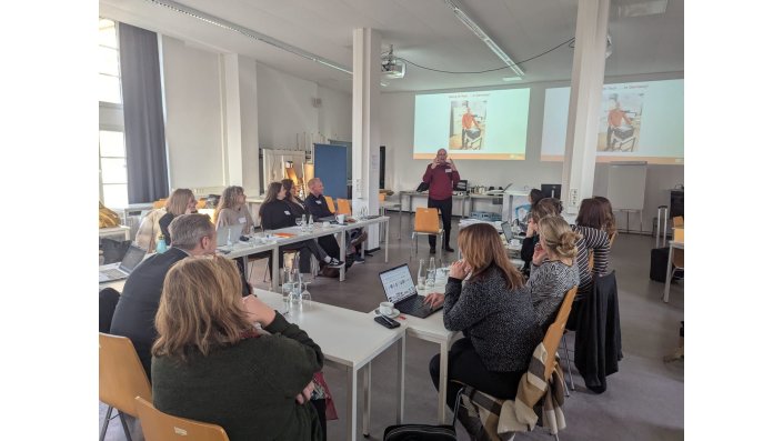 Prof. Dr. Perry Hobson wears a read sweater and black pants. He's presenting with a powerpoint in the background. In front of the picture are multiple people listening, seated at tables.