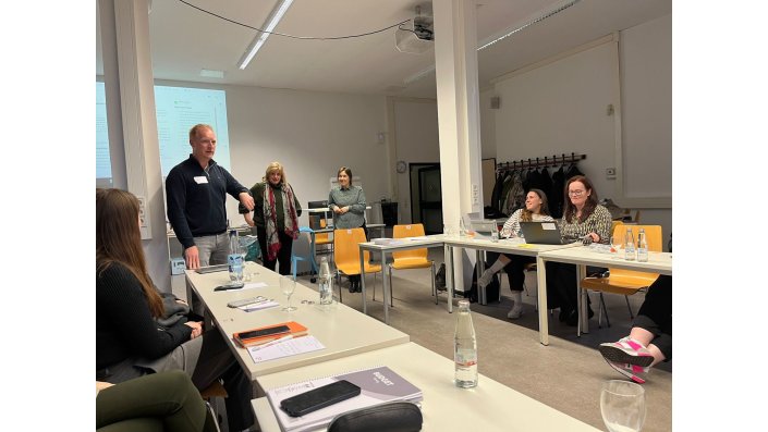 Mike Klaassen, wearing a dark blue sweater and light pants, and Susanne Glaeser, wearing a green turtleneck and red-grey scarf, are leading the seated participants in the foreground of the picture through a discussion.