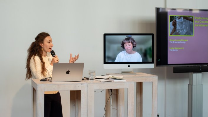 eine Frau mit Mikrofon neben einem Bildschirm, auf dem per Videokonferenz eine Frau dazugeschaltet ist
