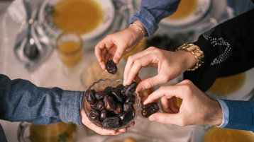 Über einem gedeckten Tisch hält eine Hand eine Schüssel mit Datteln, drei weitere Hände greifen zu und nehmen sich je eine Dattel.  (Bild: Malik Nalik/Adobe Stock)