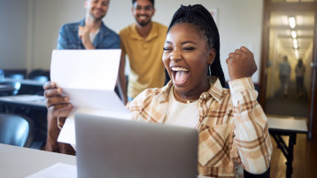 Eine Studentin, die vor einem Laptop sitzt, hält einen Brief in der Hand und freut sich über den Inhalt, sie reckt dabei die Faust nach oben in einer Siegerpose (Image: Adobe Stock / K Abrahams_peopleimages.com)